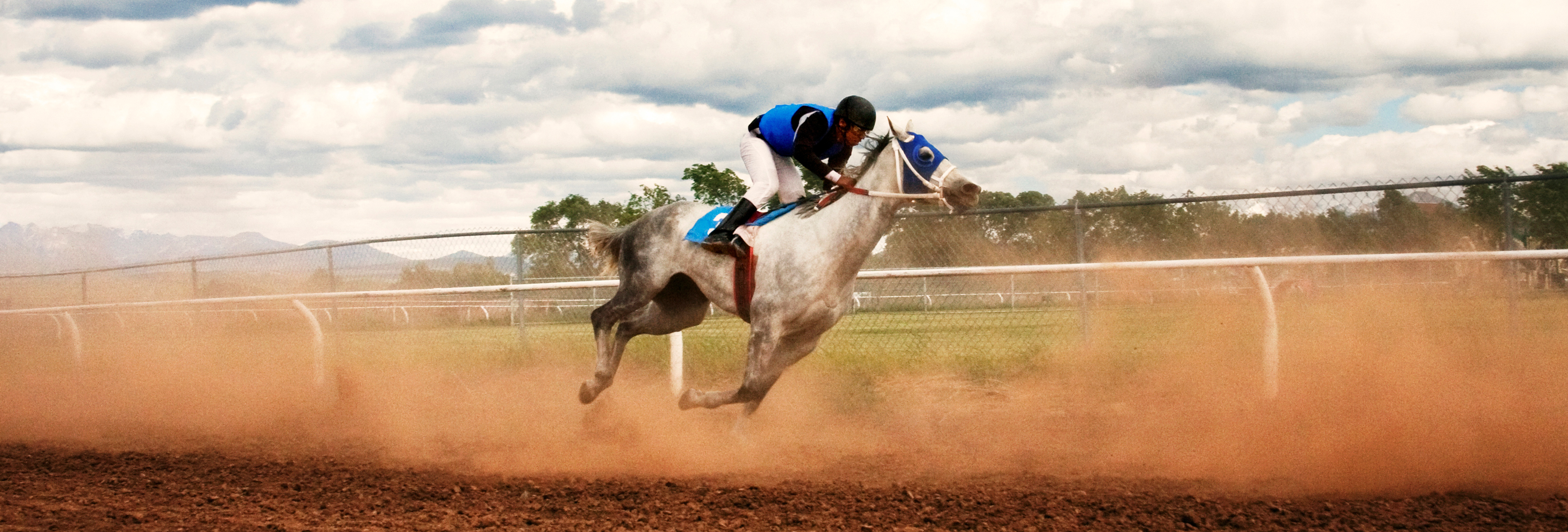 Jockey on a grey horse racing on a dirt track, kicking up dust under a cloudy sky.