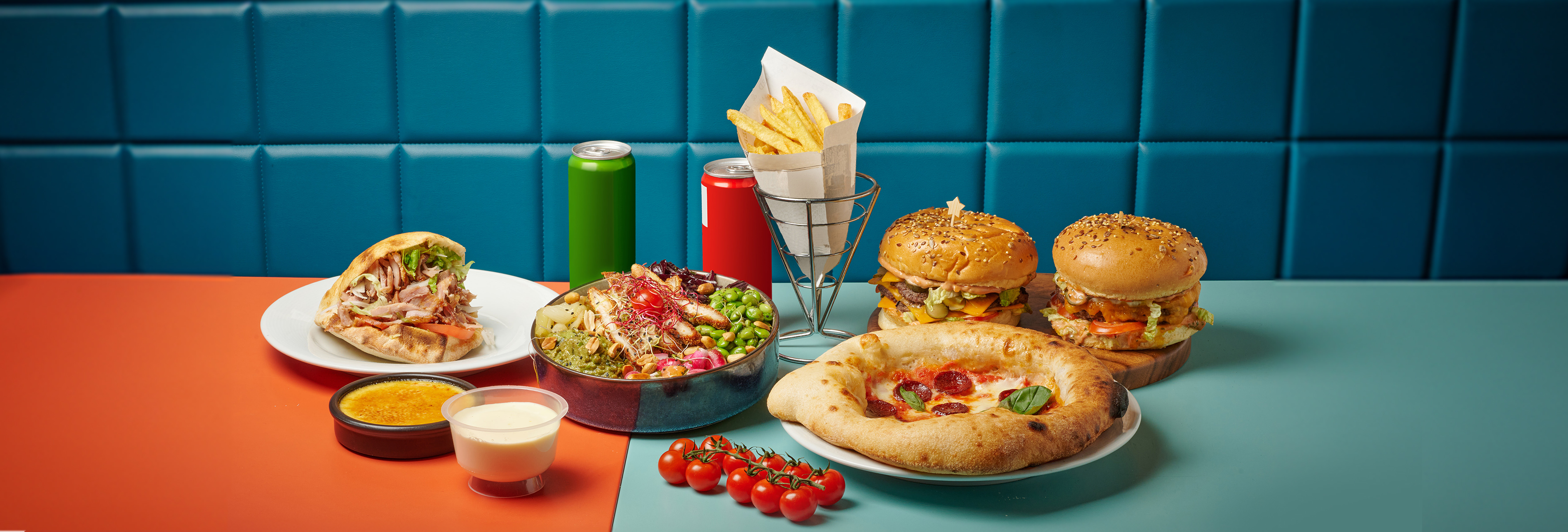 A vibrant spread of fast food sits on a two-toned table against a teal, padded wall. The food includes two burgers on a small wooden board, a personal pizza garnished with basil, a pita sandwich overflowing with meat and lettuce, a colorful salad bowl with chicken, edamame, and various vegetables, a cone of french fries in a metal holder, two cans of soda (one green, one red), a small dish of crème brûlée, and a small cup of what appears to be a creamy sauce. A small branch of cherry tomatoes rests on the teal section of the table near the pizza. The padded wall provides a textured backdrop to the scene.